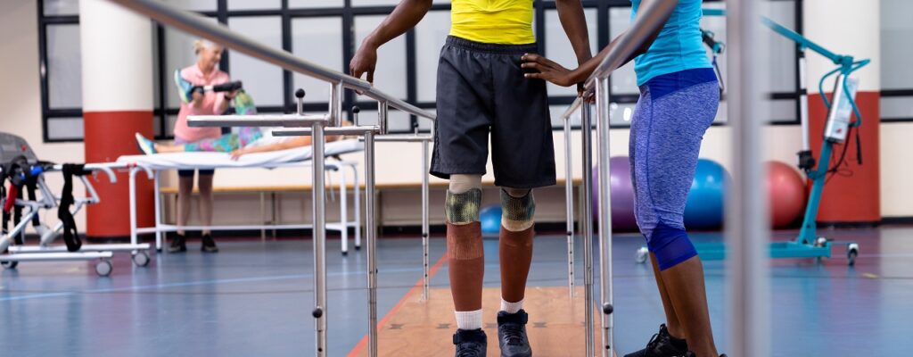 Physiotherapist helping disabled man walk with parallel bars in